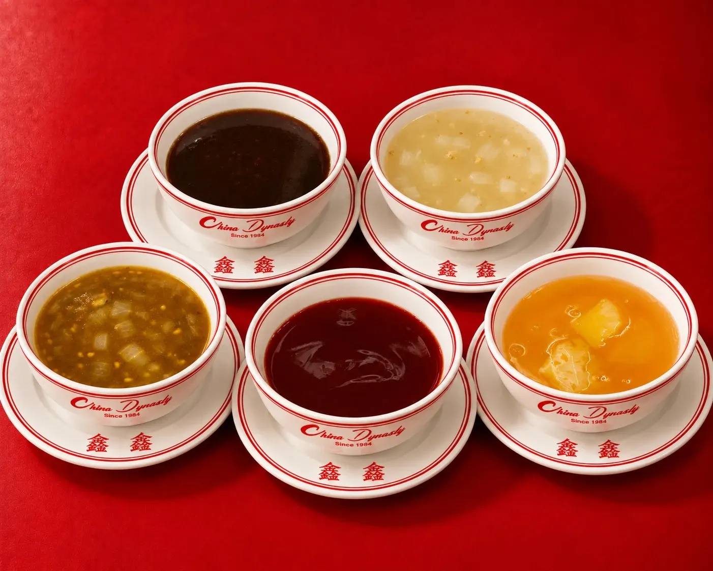 Five of Grandma’s homemade sauces in China Dynasty bowls on a red table, including brown sauce, sweet and sour, and lemon sauce with pineapple